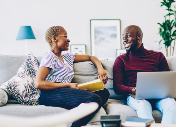 Cheerful african american marriage spending time at home enjoying free time together, positive woman sitting with book while communicating with her husband using laptop computer and wifi in apartment